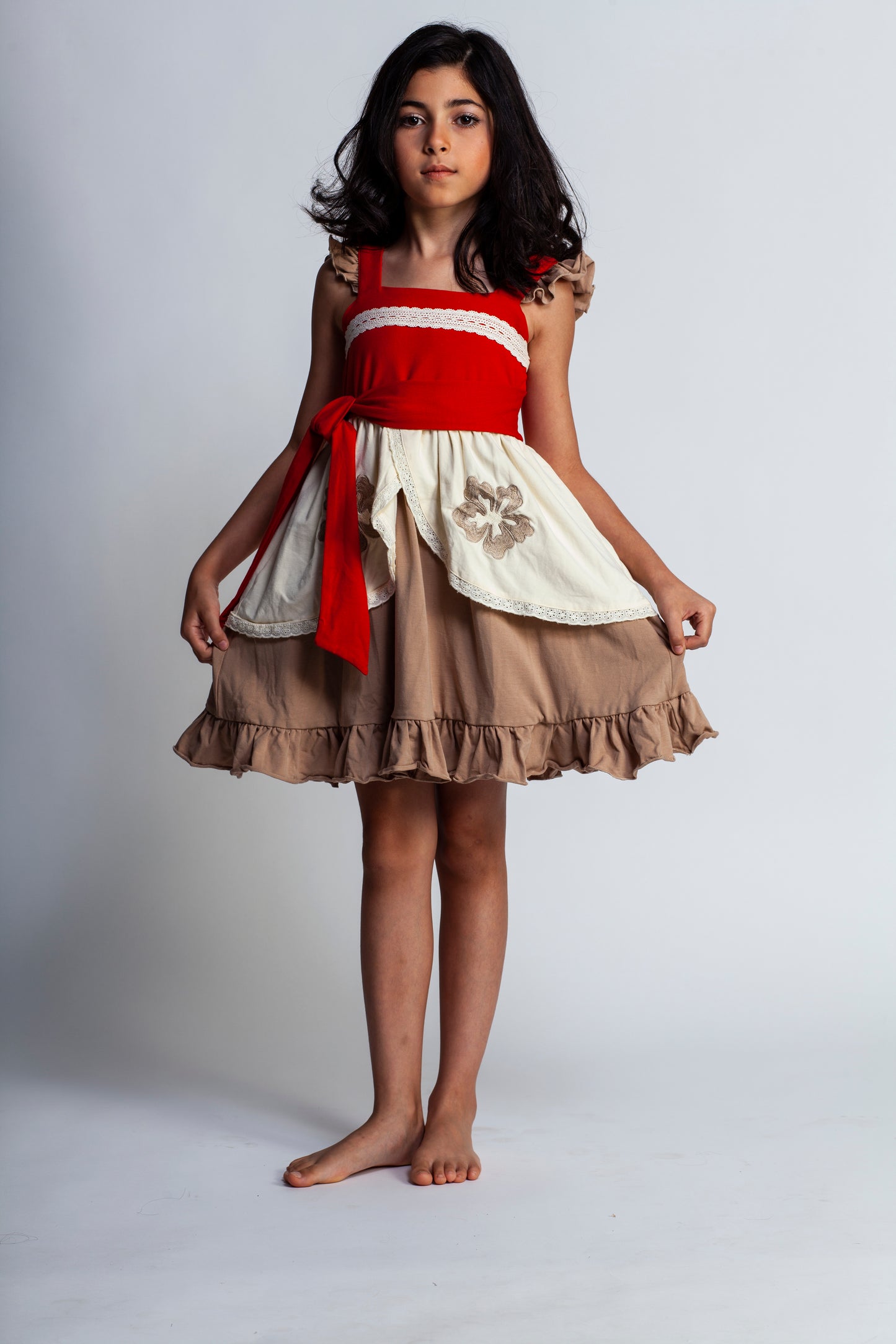 A young girl is wearing the Polynesian Princess Twirl Dress, which features beige and cream fabric accented by a red waistband and decorative floral embroidery.  There is a lace accent on the chest and ruffles on the top of the straps.  There is a ruffle on the hem and a tie on the side of the dress. Perfect for an adventurous princess. 