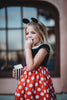 A young girl is wearing the Miss Mouse Twirl Dress, which features a black bodice, puff sleeves and a white collar.  Three white buttons decorate the center. The skirt is red with white polka dots and white eyelet lace trim.