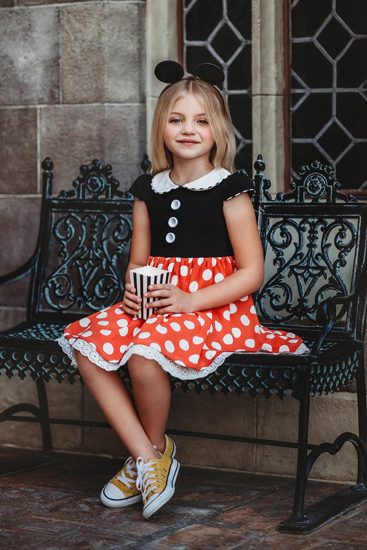 A young girl is wearing the Miss Mouse Twirl Dress, which features a black bodice, puff sleeves and a white collar.  Three white buttons decorate the center. The skirt is red with white polka dots and white eyelet lace trim.