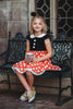 A young girl is wearing the Miss Mouse Twirl Dress, which features a black bodice, puff sleeves and a white collar.  Three white buttons decorate the center. The skirt is red with white polka dots and white eyelet lace trim.