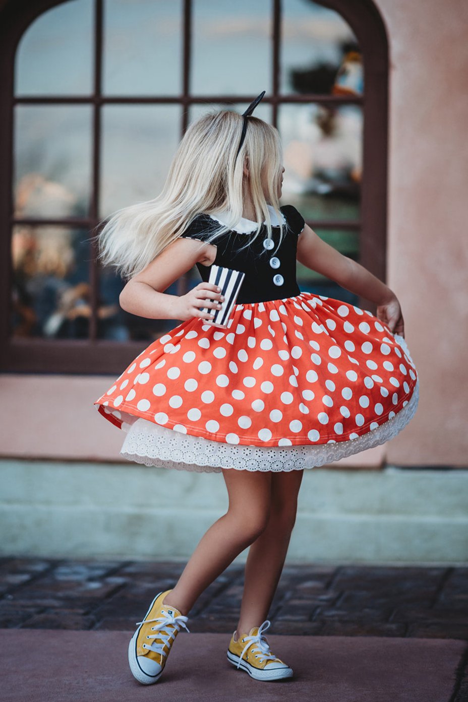 A young girl is wearing the Miss Mouse Twirl Dress, which features a black bodice, puff sleeves and a white collar.  Three white buttons decorate the center. The skirt is red with white polka dots and white eyelet lace trim.