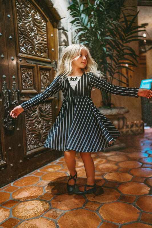 A young girl is wearing a Black and white stripe cotton twirl dress with long sleeves.  The front has a bat motif embroidered  on a white v. 