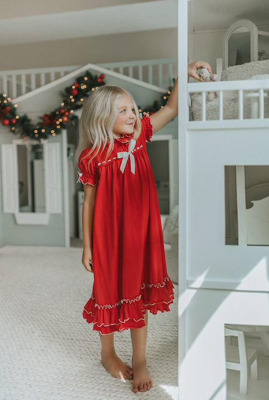 A young girl is wearing The Original Clara Nutcracker Gown in red, featuring red ruffles and a white bow. The buttery soft fabric is cozy and sensory friendly.  The puff sleeves with ribbon detail and the ruffles at the hem add a traditional classic look. 