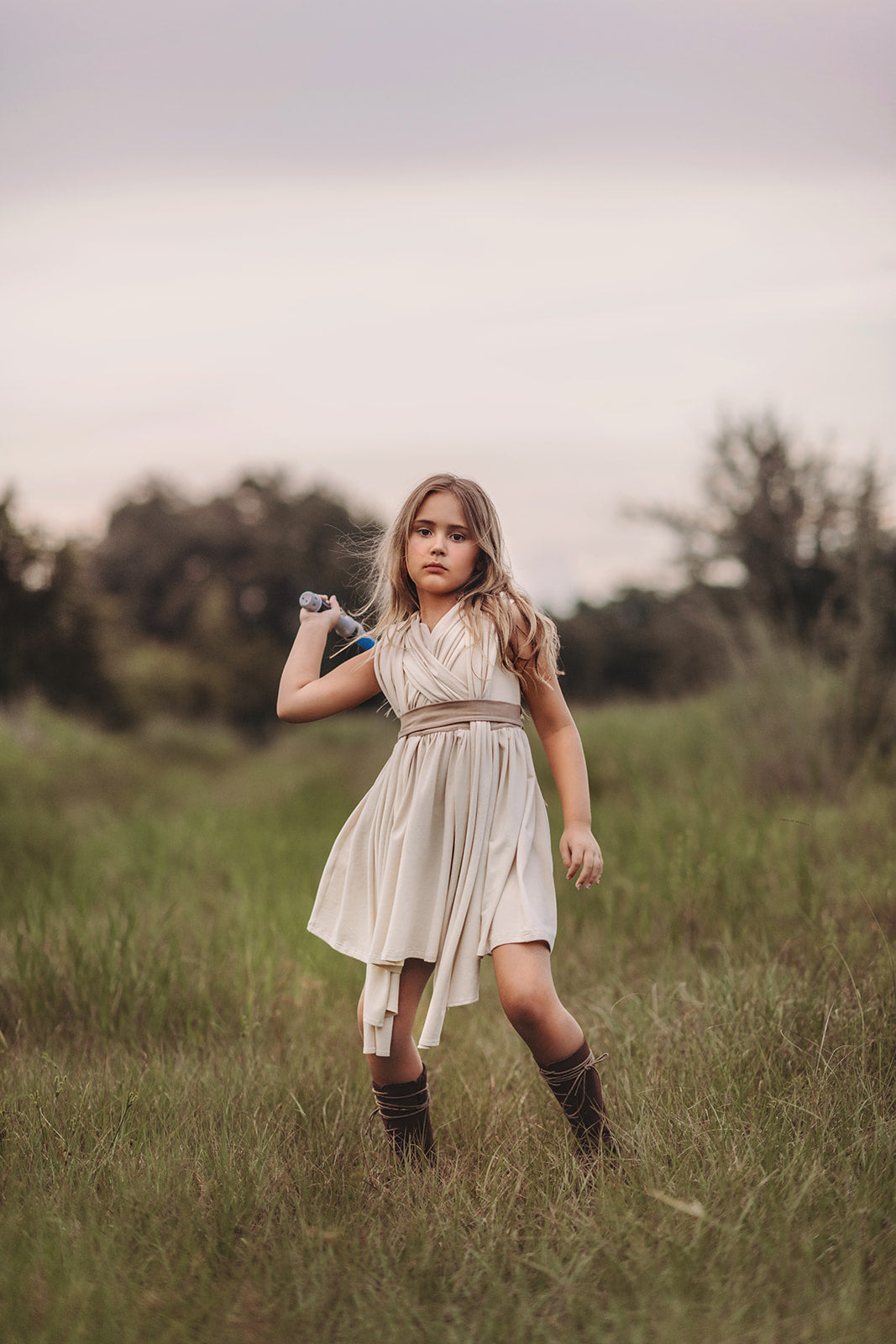 In a far away galaxy a girl and her lightsaber stand in a beige dress.  There are layers of fabric that cross and intersect on the bodice.  The cotton twirl dress ties at the waist in the back. 