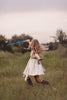 In a far away galaxy a girl and her lightsaber stand in a beige dress.  There are layers of fabric that cross and intersect on the bodice.  The cotton twirl dress ties at the waist in the back. 