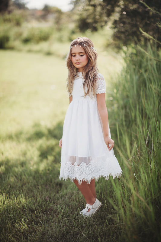 A young girl with long wavy hair, wearing a Victorian Day Gown in White and a floral headband, stands gracefully in a sunlit grassy field. She looks down, holding the hem of her elegant dress with one hand. The background is blurred, with tall green grass and trees visible.