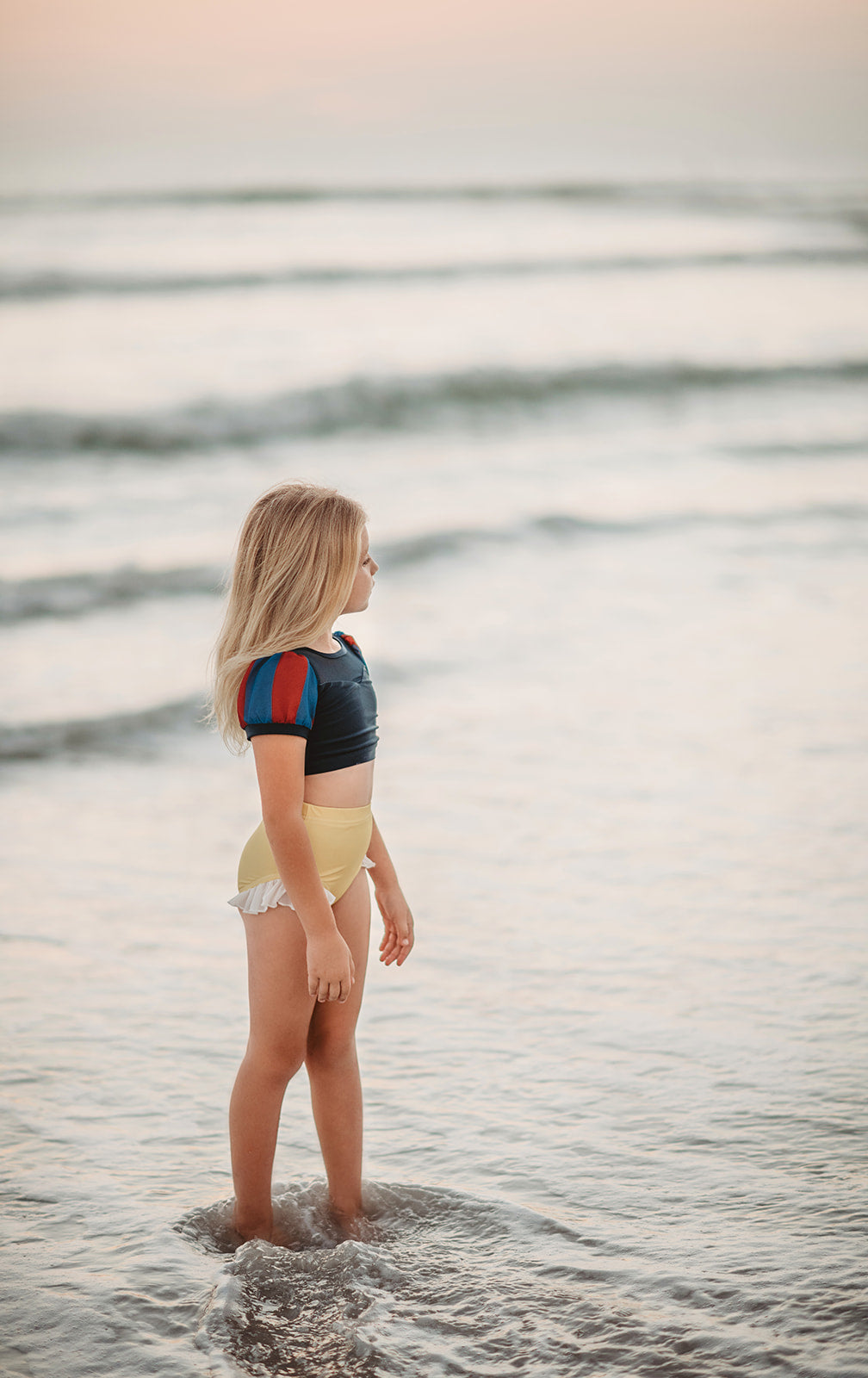 A young girl is wearing the Snow White  2 piece Swim Set with SPF 50 for sun protection.  The navy blue top has blue and red puff sleeves and navy mesh at the top.  The light yellow bottoms have a white ruffle on the leg. 