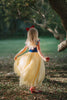 A young girl stands in a forest wearing the Portrait Collection Snow White Gown. The gown is velvet blue and a light yellow skirt with a red ribbon criss cross accent on the bodice. The dress is sleeveless with a red and blue flutter ruffle on the top. 