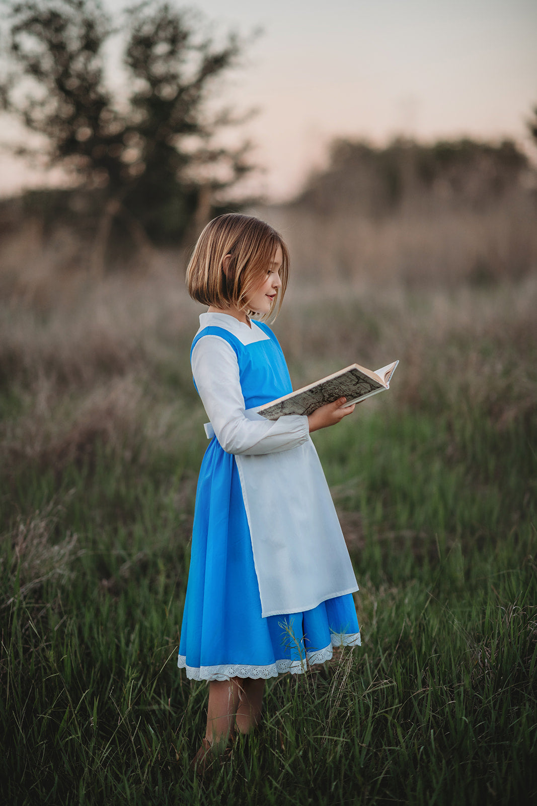 A young girl stands wearing the Red Rose Princess Village Dress from the Fairy Godmother Collection, reminiscent of Disney's Belle from "Beauty and the Beast." The white sleeves are long. The dress has a bight blue chest and skirt.  There is a white apron at the waist and white embroidery lace at the base. Dress is mid calf.
her 100% woven cotton dress to flare out. The background is softly blurred with trees and sky visible.