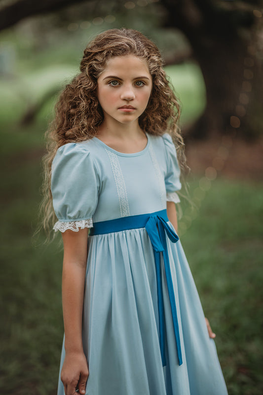 A young girl in the Lost Child Blue Gown with puffed sleeves and a blue ribbon tied at the waist. There is lace at the bottom of the sleeves and in two vertical stripes on the chest. 