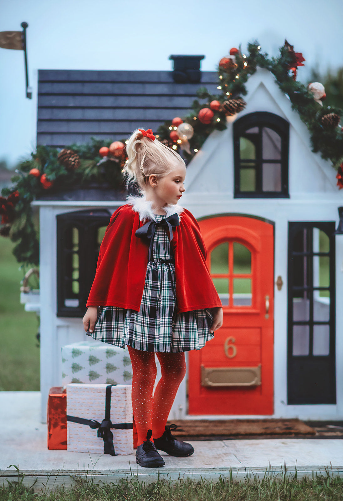 A young girl stands dressed in a black and white vintage plaid dress. A red velvet cape trimmed with white fur and a black bow are tied around her shoulders. The dress is mid thigh length and a sewn in pettiskirt add fullness to the bottom of the dress. 