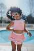 A young girl is wearing a 2 piece pink swim set. There is white collar with a pink bow at the neck.  Two lighter pink stripes are on the chest like ribbon tails.  The bottom is a skirt with bows light pink bows at the hem.