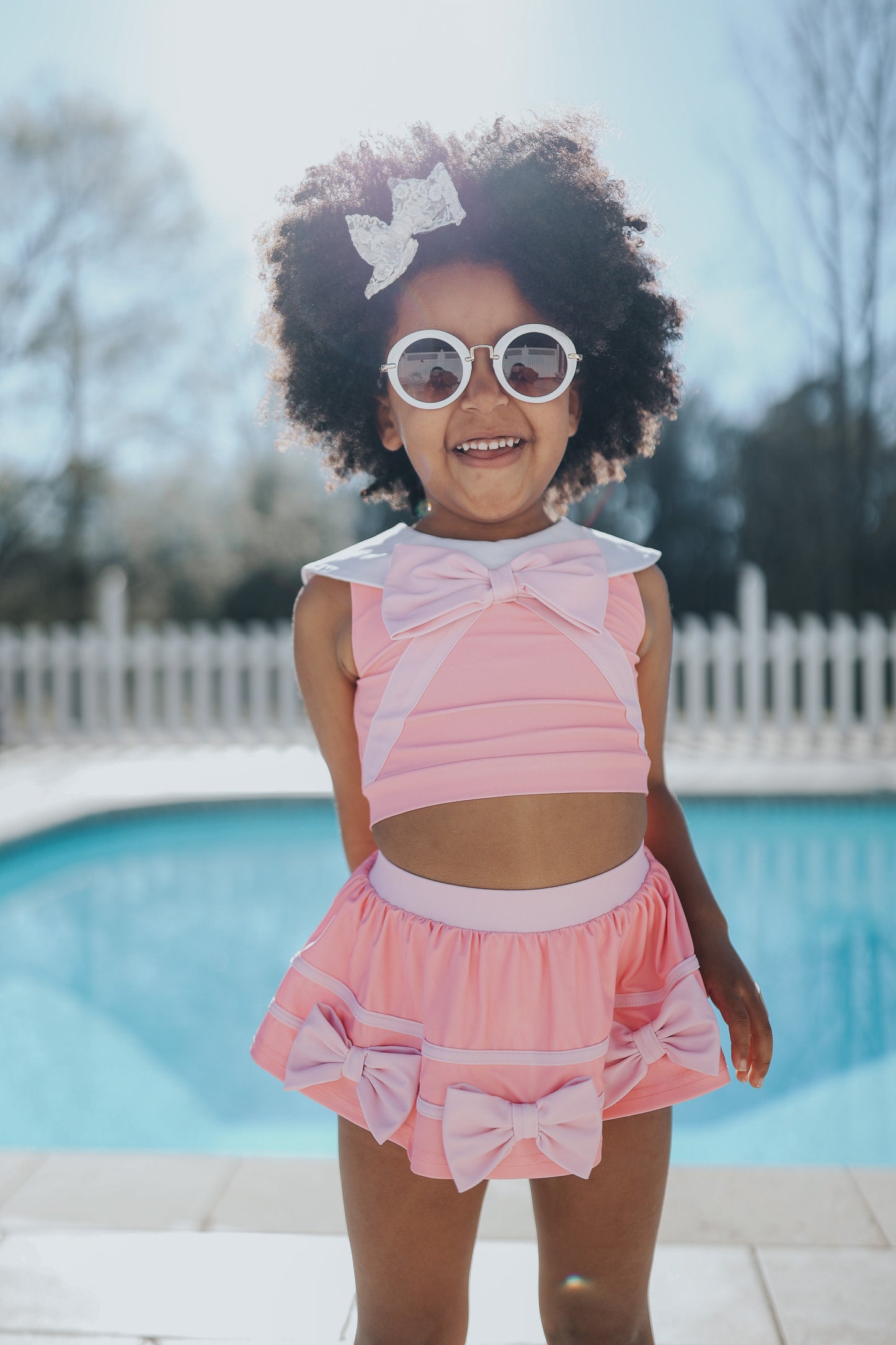 A young girl is wearing a 2 piece pink swim set. There is white collar with a pink bow at the neck.  Two lighter pink stripes are on the chest like ribbon tails.  The bottom is a skirt with bows light pink bows at the hem.