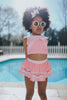 A young girl is wearing a 2 piece pink swim set. There is white collar with a pink bow at the neck.  Two lighter pink stripes are on the chest like ribbon tails.  The bottom is a skirt with bows light pink bows at the hem.