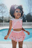 A young girl is wearing a 2 piece pink swim set. There is white collar with a pink bow at the neck.  Two lighter pink stripes are on the chest like ribbon tails.  The bottom is a skirt with bows light pink bows at the hem.