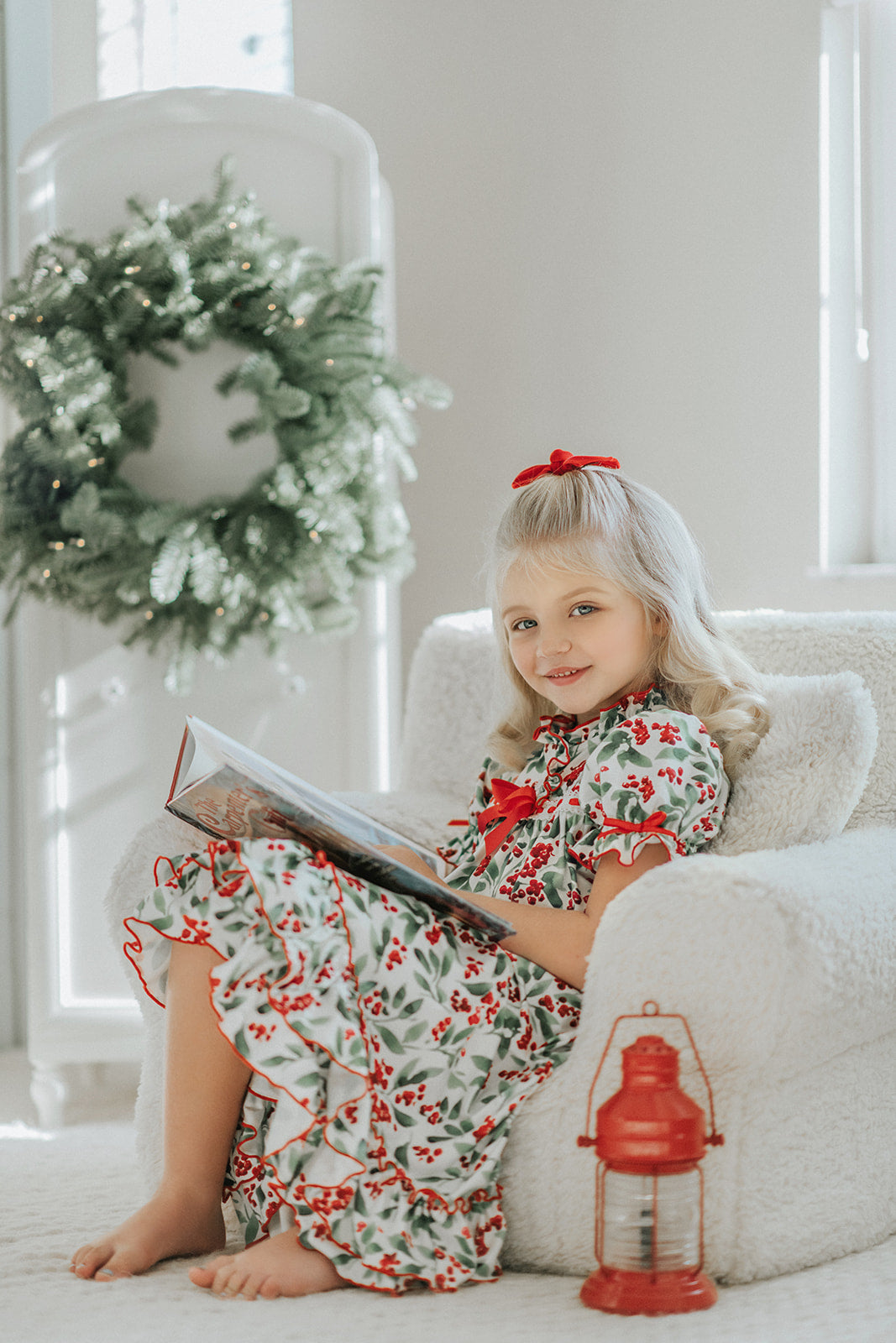 A young child with curly blonde hair stands barefoot on a soft carpet, holding an open book. The child wears *The Original Clara Nutcracker Gown in Winterberry* and reads intently. A decorated Christmas tree is visible in the background, part of our exclusive Christmas Lounge Collection.