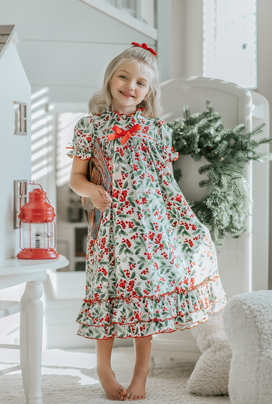 A young child with curly blonde hair stands barefoot on a soft carpet, holding an open book. The child wears *The Original Clara Nutcracker Gown in Winterberry* and reads intently. A decorated Christmas tree is visible in the background, part of our exclusive Christmas Lounge Collection.