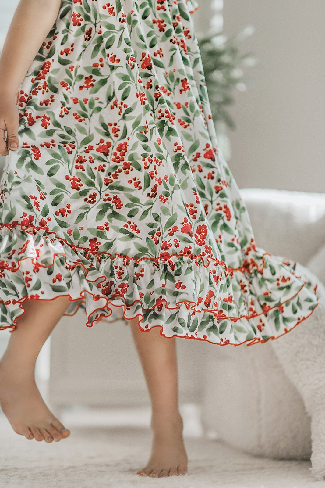 A young child with curly blonde hair stands barefoot on a soft carpet, holding an open book. The child wears *The Original Clara Nutcracker Gown in Winterberry* and reads intently. A decorated Christmas tree is visible in the background, part of our exclusive Christmas Lounge Collection.