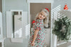 A young child with curly blonde hair stands barefoot on a soft carpet, holding an open book. The child wears *The Original Clara Nutcracker Gown in Winterberry* and reads intently. A decorated Christmas tree is visible in the background, part of our exclusive Christmas Lounge Collection.