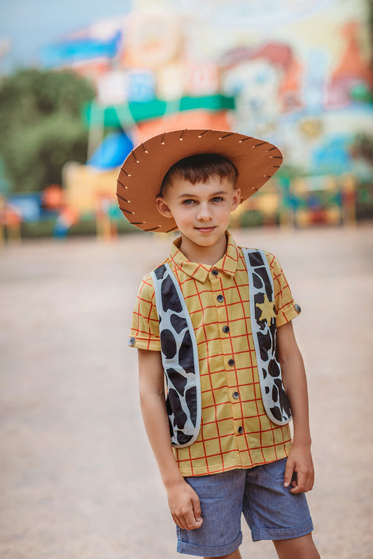 A young boy stands dressed in a sheriff cowboy outfit from the Toyland Collection. A yellow and red checkered short sleeve shirt featuring black and white cow print vest. A yellow sheriff badge is sewn on the right chest of the vest. The collar shirt features buttons in the middle and one on each sleeve.